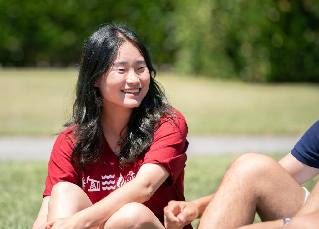 A student sits on the grass and smiles at someone sitting next to her whose face is out of frame.